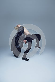 Shirtless man in black pants and heeled boots bending backward in artistic dance move against grey studio background.