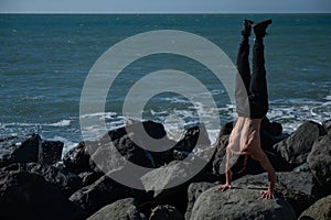 Shirtless man doing handstand on rocks by the sea.