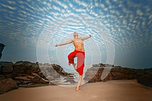 A shirtless man in red pants jumps joyfully against a dramatic sky, with rocky cliffs and a sandy beach below, exuding a sense of