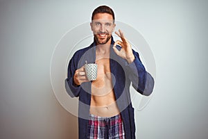 Shirtless man wearing comfortable pajamas and robe drinking cup of coffee doing ok sign with fingers, excellent symbol
