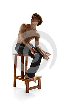 Shirtless muscled man with naked torso posing sitting on stool against white studio background. Male model with curly