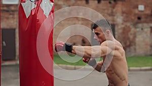 Shirtless muscular boxer punching a punching bag while training outdoors. Close-up