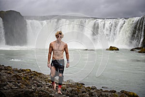 Shirtless and muscular boy walks around the Godafoss waterfall in Iceland