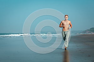 A shirtless, muscular man jogs along the wet sand of a beach, with the ocean and mountains in the background. He has an intense,