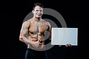 Shirtless muscular young man standing holding a blank horizontal white banner pointing finger at the copyspace for your