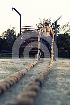 Shirtless ripped muscular man wearing running mask for respiratory boost working out with a battle rope outdoors