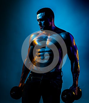 Shirtless sexy bodybuilder is doing exercise with dumbbell. Neon blue light.