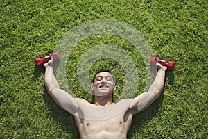 Shirtless, smiling, muscular man lying in grass with dumbbells in Beijing, view from above