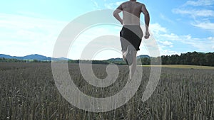 Shirtless sportsman running over harvested field. Legs of athlete with black sneakers run in nature at sunset.