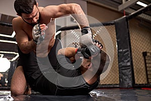 shirtless strong mma fighter in boxing gloves sitting above opponent while sportsman lying on floor resisting