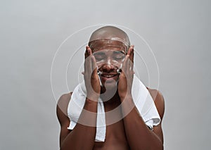Shirtless young african american man smiling while washing his face with foam cleanser, standing with towel around his