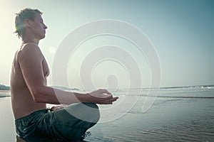 A shirtless young man sits in a meditative lotus pose on the wet sand of a beach, with the ocean waves crashing behind him