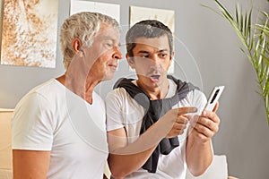 Shocked adult man sitting with his senior father in living room at home using smartphone see shock content browsing internet web