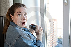 Shocked adult woman after spying out the window with binoculars, domestic room