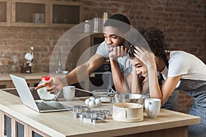 Shocked african-american couple baking with recipe on laptop