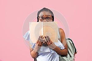 Shocked African American female student with backpack hiding behind open book on pink studio background