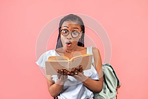 Shocked African American female student with backpack staring into open textbook on pink studio background