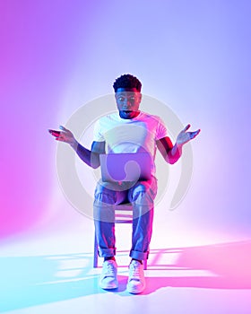 Shocked African American guy looking at laptop screen, opening mouth in surprise, sitting on chair in neon light