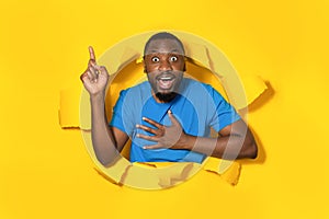 Shocked african american man pointing up at free space through hole in ripped yellow paper, banner design