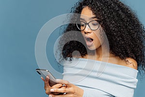 Shocked African American woman gots message from ex, gasps from surprise and excitement, reads stunning news on mobile phone, has