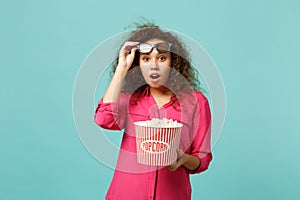Shocked african girl in 3d imax glasses watching movie film and holding popcorn isolated on blue turquoise wall