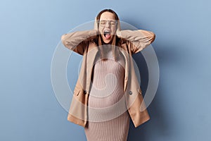 Shocked angry stressed pregnant woman in beautiful dress and jacket posing  over blue background covering ears with hands