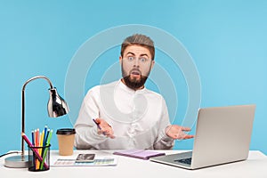 Shocked annoyed man office worker spreading hands looking at camera with astonishment, sitting at workplace with laptop and coffee