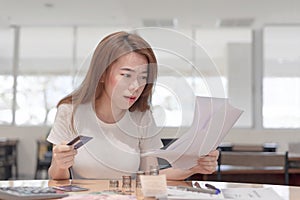 Shocked asian business woman holding credit card sitting on desk looking at bills payment in office background