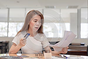 Shocked asian business woman holding credit card sitting on desk looking at bills payment in office background