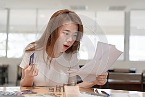 Shocked asian business woman holding credit card sitting on desk looking at bills payment in office background