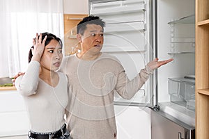 Shocked asian couple standing next to empty fridge at home