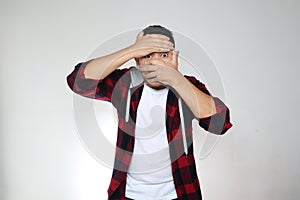 Shocked Asian man covering his face, peeking trough fingers. Worried afraid expression over white