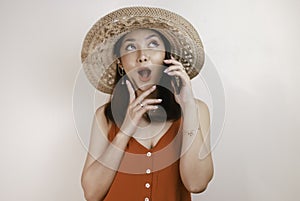 Shocked Asian woman wearing a straw hat talking on a mobile phone and laughing, expressing excitement emotions having a pleasant