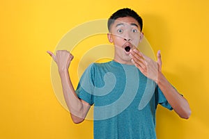 Shocked asian young man pointing a copy space with thumb opening his eyes very wide covering his mouth with hand