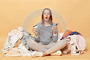 Shocked astonished Caucasian woman posing near heap of multicolored unsorted clothes isolated over beige background looking at