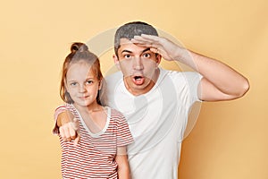 Shocked astonished man looking far with hand near forehead and little kid girl pointing to camera at you standing together