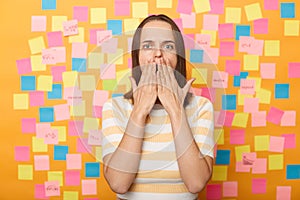 Shocked astonished surprised amazed woman wearing striped t shirt, covering widely open mouth with palms, standing isolated over