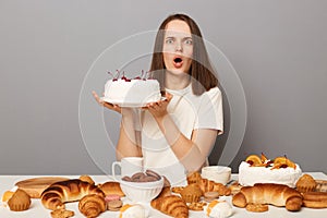 Shocked astonished surprised woman wearing white T-shirt isolated over gray background holding big cake sitting at festive table