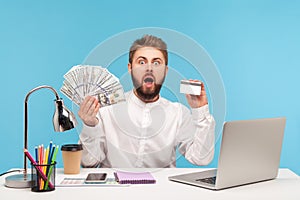 Shocked bearded man office worker holding dollar cash and credit card looking at camera with astonishment, surprised with high