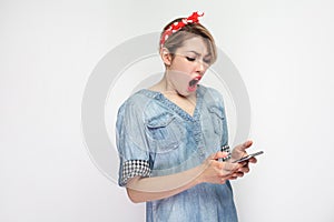 Shocked beautiful blogger young woman in casual blue denim shirt with makeup,red headband standing, use smartphone and watching