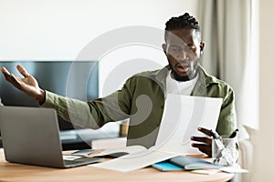 Shocked black man reading paper letter mail and spreading hands, sitting at table with laptop at home office