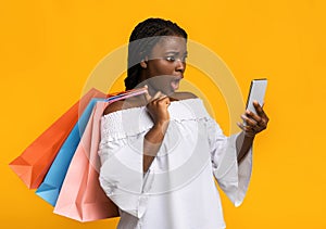 Shocked black shopaholic lady looking at smartphone and holding lots shopping bags