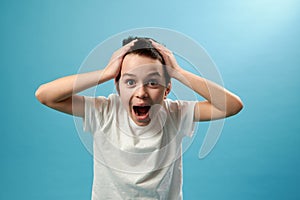 Shocked boy screams and laughs with his hands on his temples, expressing shock and surprise. Blue background, copy space