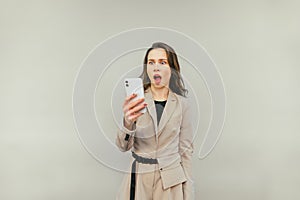 Shocked business woman looking at smartphone screen with surprised face on beige background. Isolated