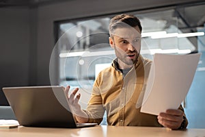 Shocked Businessman Reading Papers Having Problem Using Laptop In Office