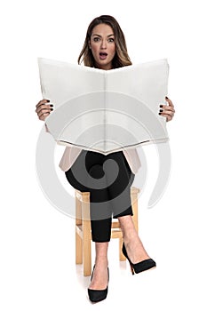 Shocked businesswoman sits on wooden stool while holding newspaper