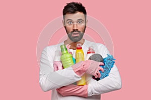 Shocked Caucasian unshaven man carries cleaning supplies, sponge for washing dish, dressed in casual outfit, stares at