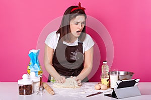 Shocked Caucasian women baking with recipe on tablet, stand with opened mouth, has surprised facial expession, kneads dough,