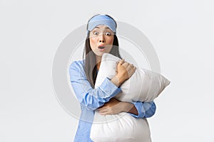 Shocked and concerned young asian girl in sleeping mask and pyjama, hugging pillow and looking tensed as watching