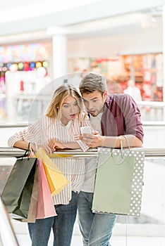 shocked couple of shoppers with paper bags looking at smartphone in shopping